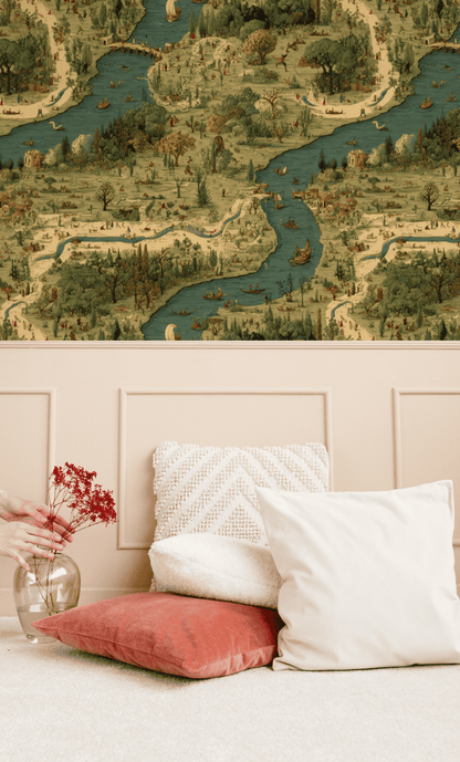 A beige wall adorned with the Pastoral Journey Wallpaper depicting rivers and villages hovers above a cushioned bench. On the floor, cushions and a glass jar with red dried flowers create a charming pastoral scene.