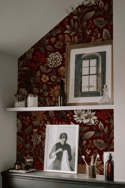 Against the Crimson Bloom Wallpaper, a shelf holds bottles, jars, and framed art, featuring a woman’s portrait and a window illustration.