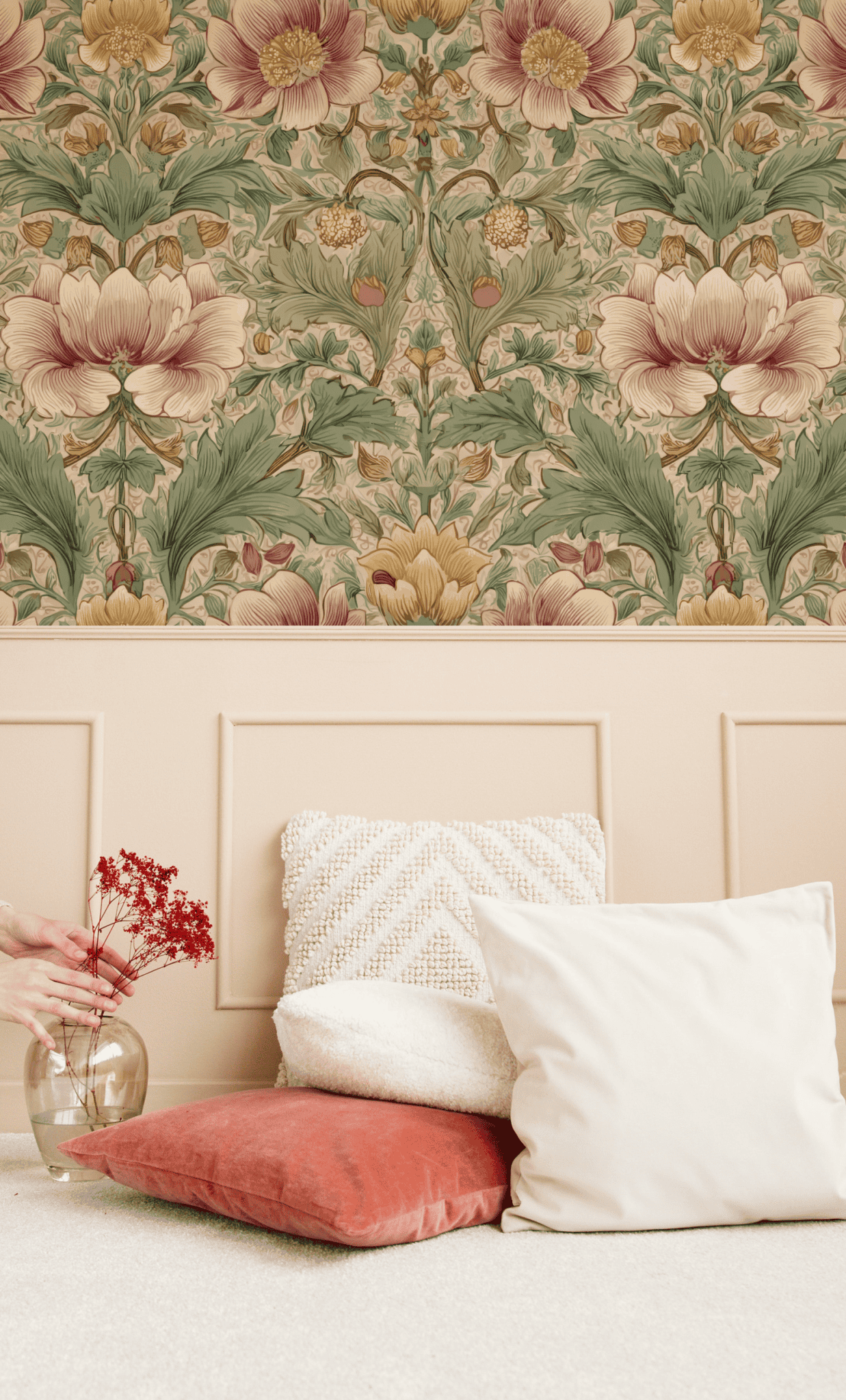 A hand places a vase with red dried flowers beside white and orange cushions on the floor; the Floral Reverie Wallpaper's floral motifs and beige wainscoting in the background evoke a timeless ambiance.