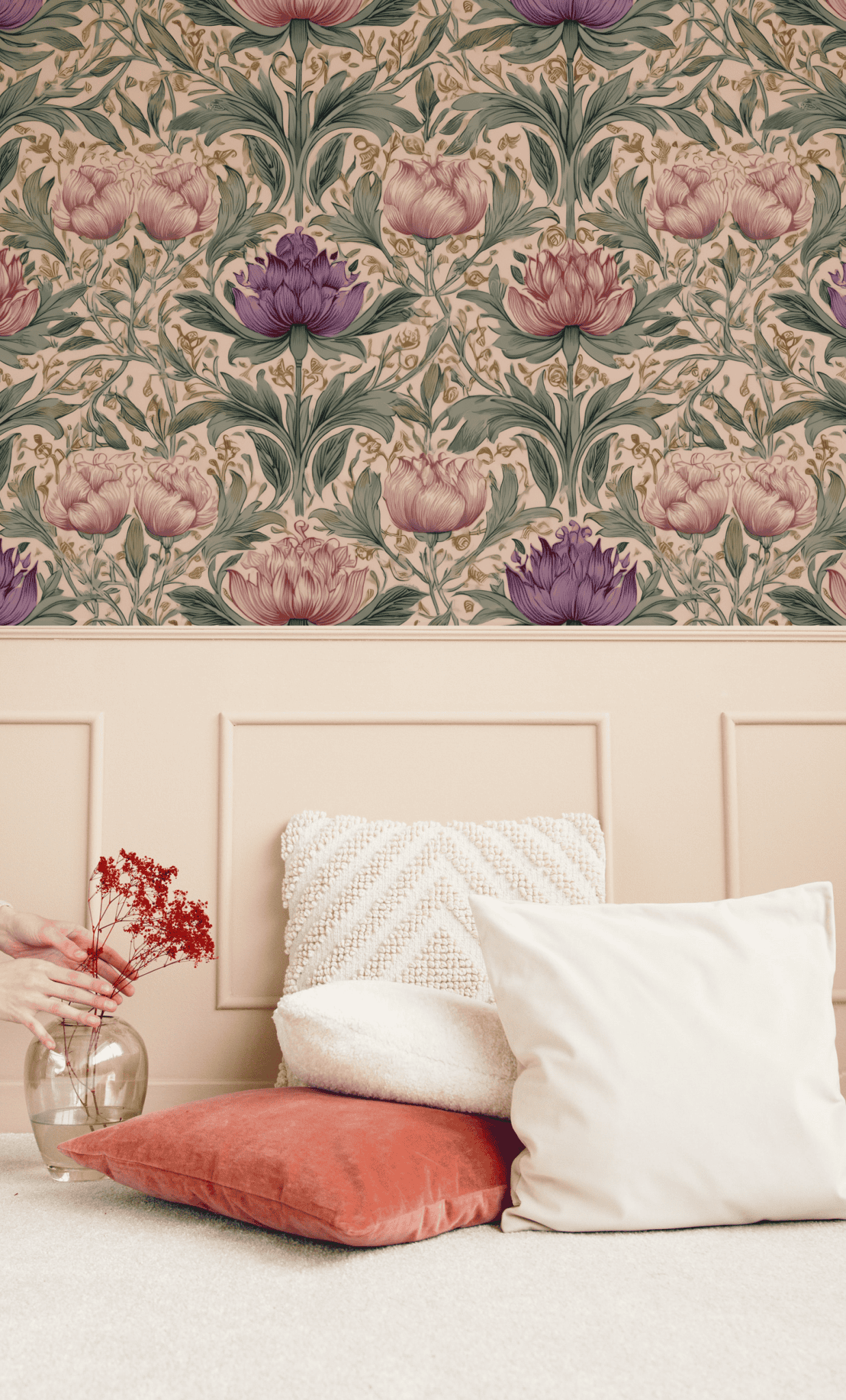 Elara Bloom Wallpaper features intricate pink and rich purple florals, paired with beige wainscoting and a stylish seating area with white and coral pillows. A hand adds elegance by placing a vase of red flowers on the floor.