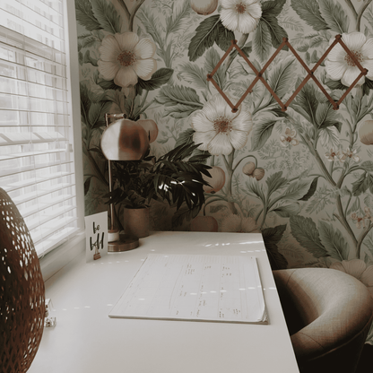 A white desk with a calendar, lamp, plant, and “be bold” card sits by a window; the wall features Eden's Bloom Wallpaper for botanical charm, complemented by a wooden accordion rack.