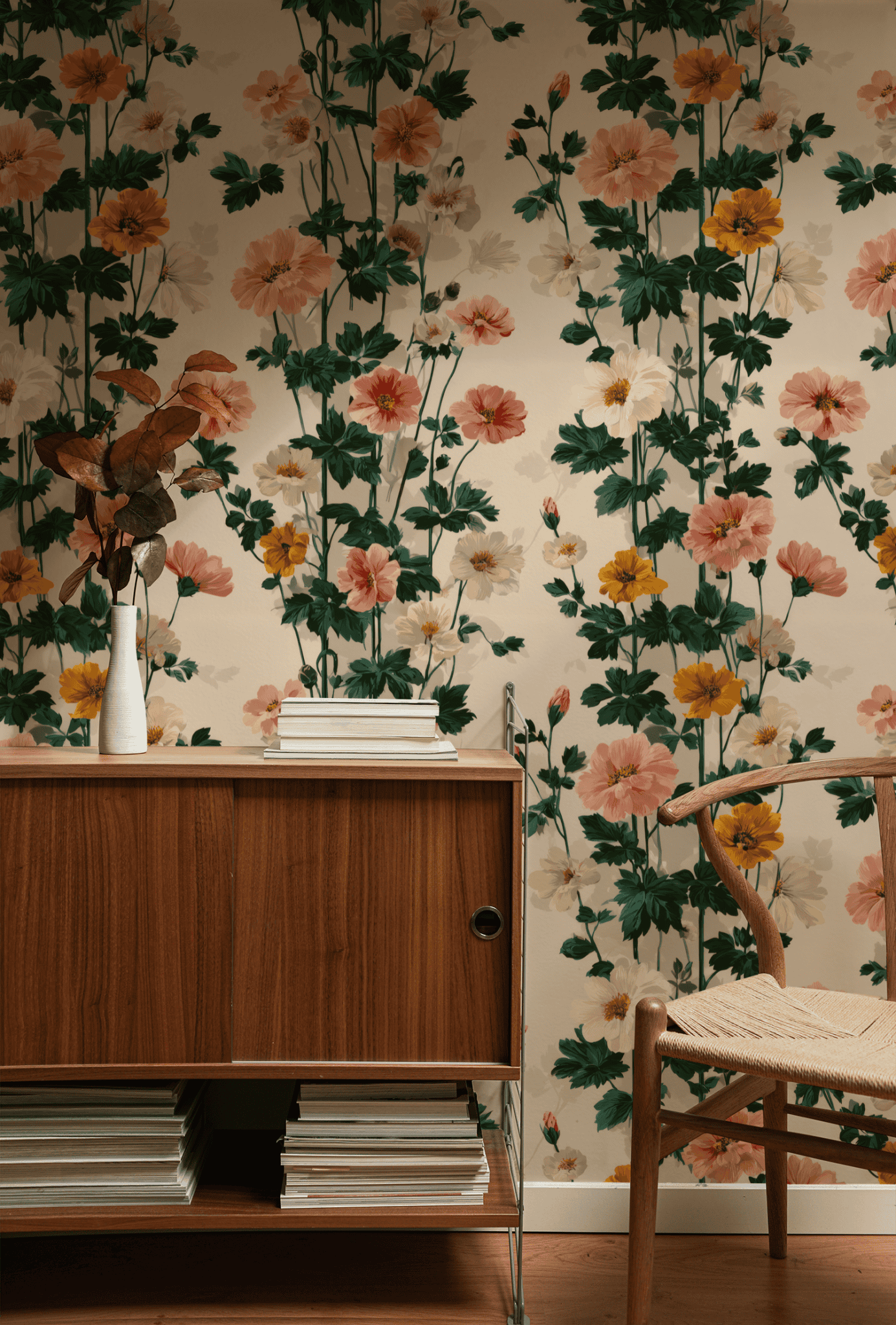 A wooden sideboard with stacked magazines and a vase of dried leaves stands in front of elegant Blossom Whisper Wallpaper, next to a woven seat chair.