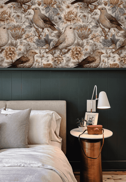 A bedroom features beige upholstery, white linens, a side table with books, a lamp, and nostalgic decor highlighted by Lark Serenade Wallpaper with bird and flower art above dark green wainscoting.