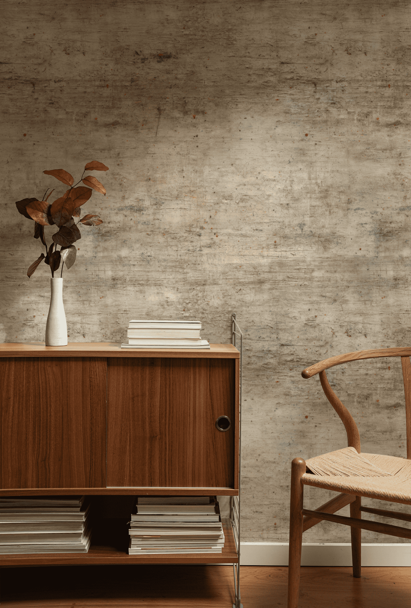 A wooden cabinet with books and a white vase of brown leaves sits by a wooden chair against Rustic Canvas Wallpaper, its earthy tones creating a cozy ambiance.