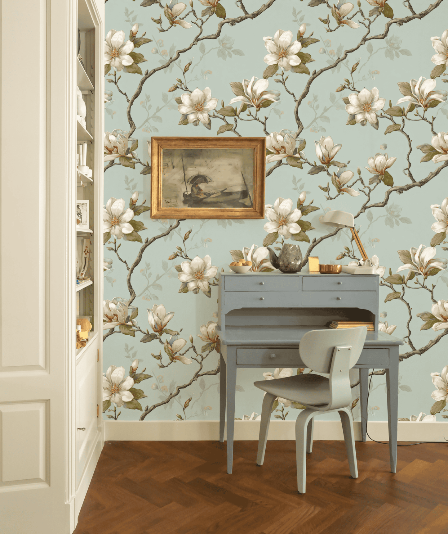 A small blue desk and chair set against a wall featuring Magnolia Grace Wallpaper exudes natural charm, with a framed painting, lamp, and decorative items on the desk beside built-in shelves.