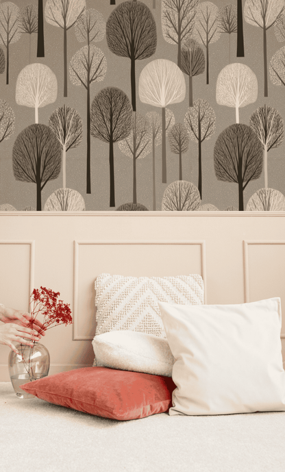 A hand arranges red flowers in a clear vase on a beige surface next to white and coral pillows, with ethereal beauty enhanced by the Winter Grove Wallpaper, its bare branches evoking a serene winter forest.