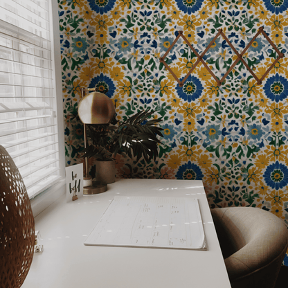 A desk with a chair, lamp, plant, notepad, and “be still” card sits by a window; behind it is vibrant decor featuring Floral Harmony Wallpaper and a geometric wall shelf.