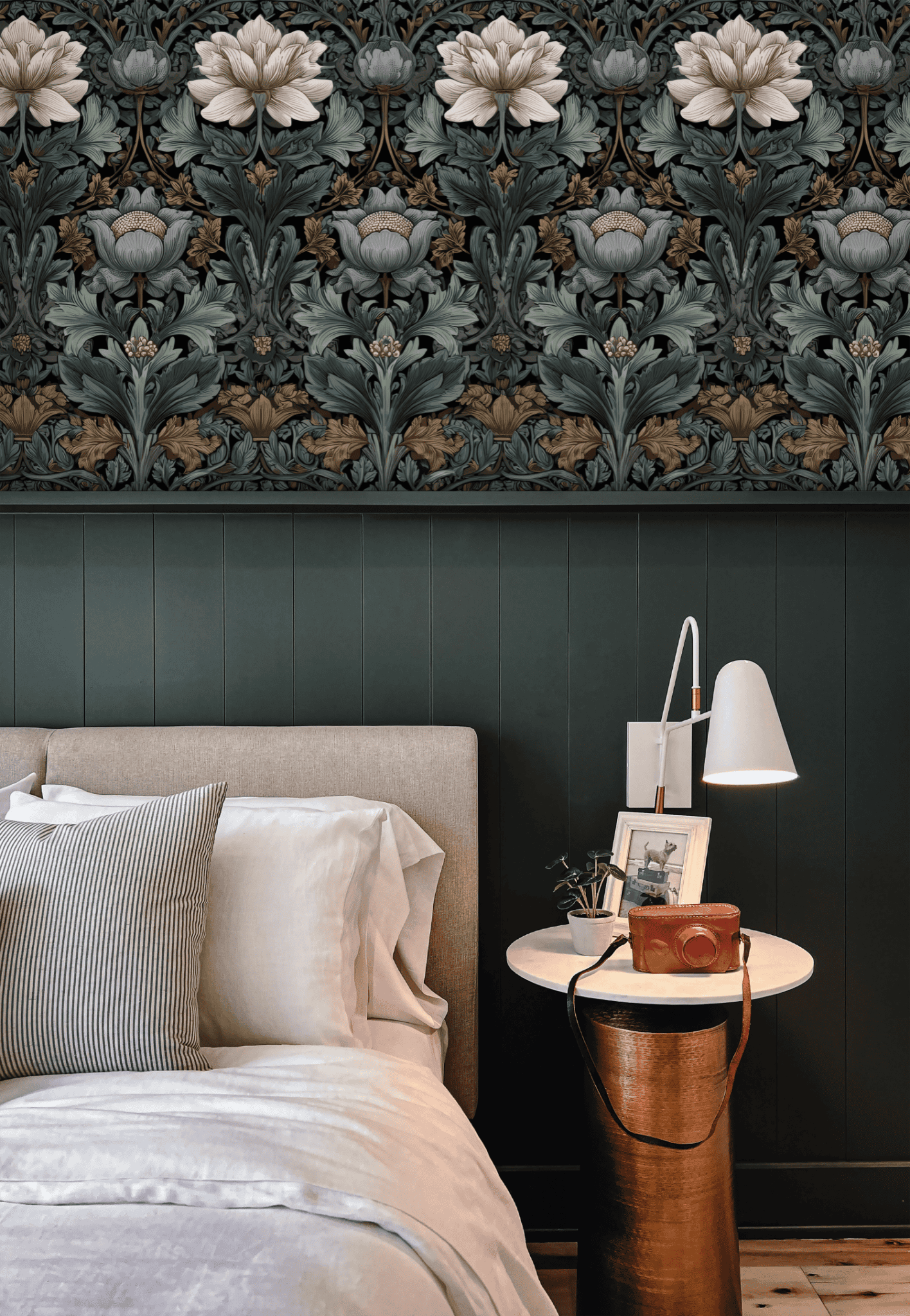 A bedroom with timeless elegance features a beige upholstered bed, white bedding, a white lamp on a round nightstand, and Vintage Blooms Wallpaper above dark green wainscoting.