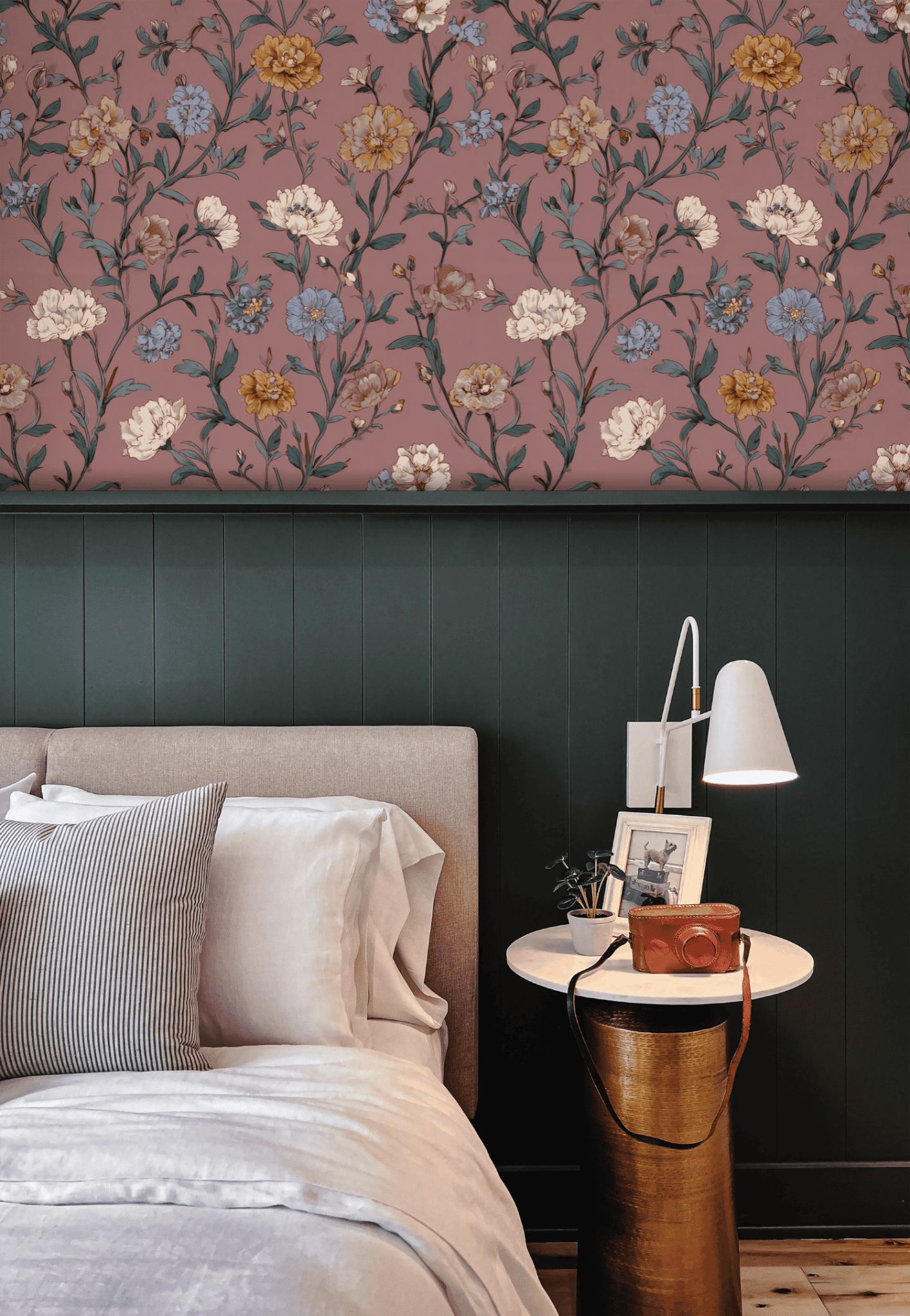 A bed with neutral bedding beside a round side table and lamp is set against dark green paneling and Blossom Reverie Wallpaper, featuring pink, yellow, and blue florals that evoke romantic tranquility with delicate blooms.