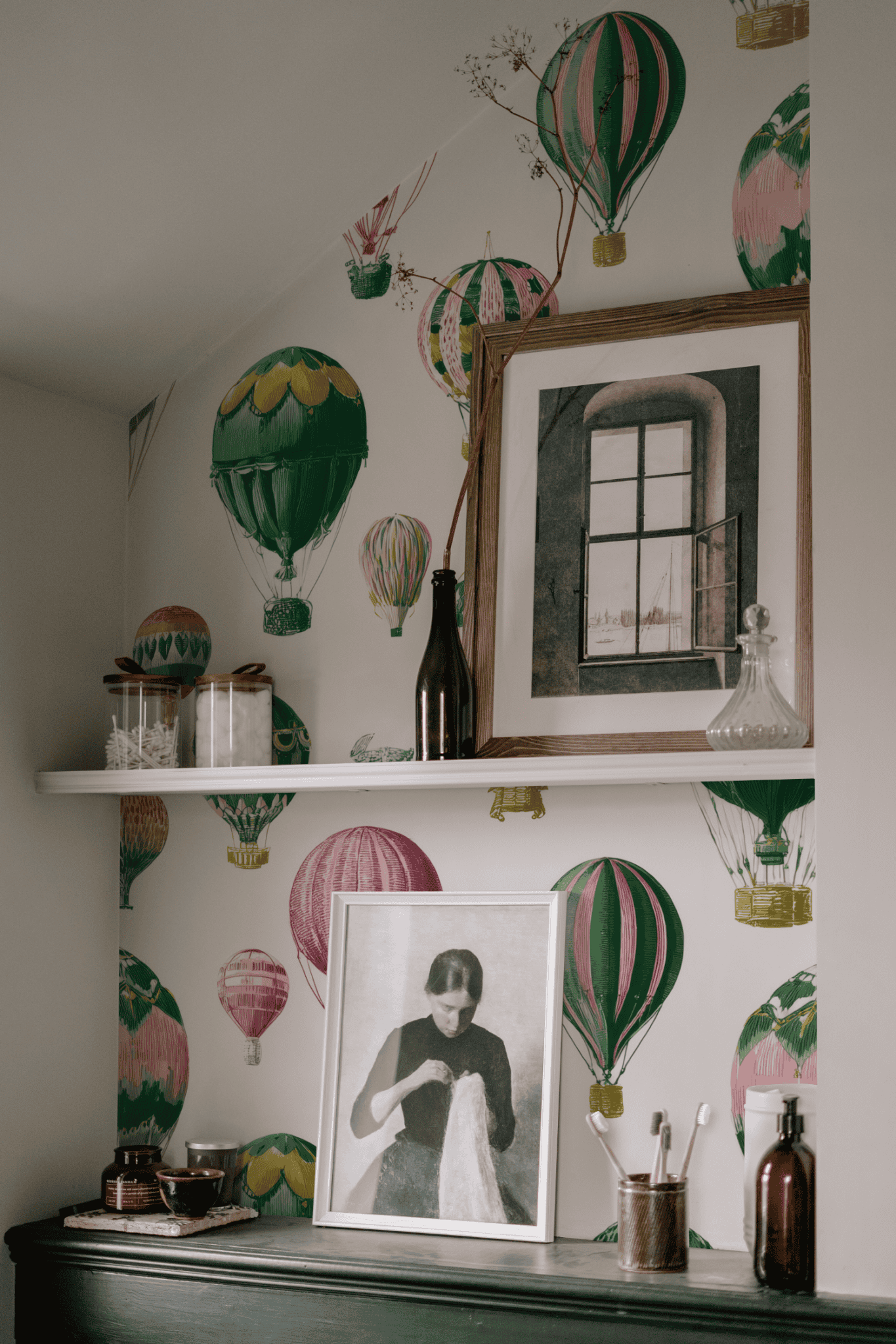 A shelf with bottles, a framed window photo, and glass containers is mounted on a wall covered in Celestial Voyage Wallpaper, adding adventurous charm; below is a black-and-white portrait of a woman in a frame.