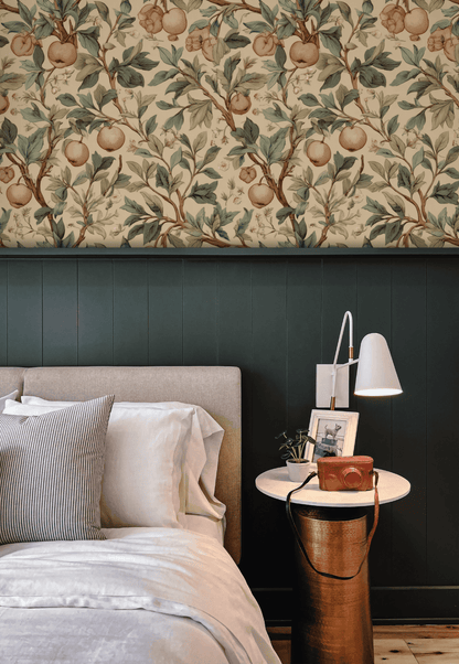 A bed with neutral bedding sits by a round white nightstand, lamp, and decor, set against dark paneling and Orchard Elegance Wallpaper showcasing fruit-laden branches for a vintage botanical touch.