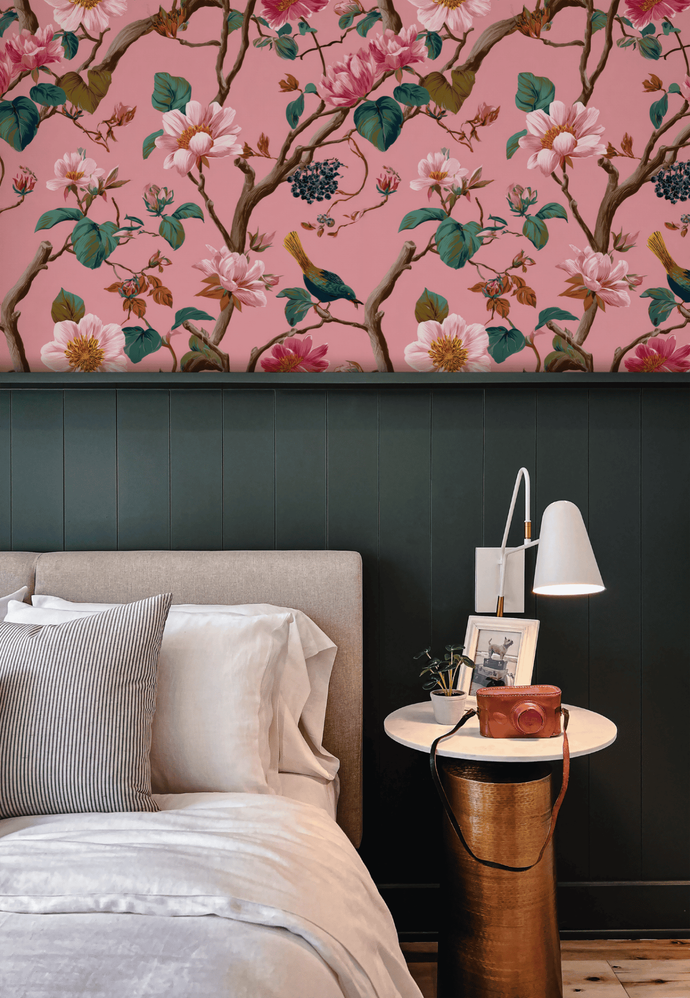 A bed with white and gray bedding sits beside a round nightstand with a white lamp, set against a wall decorated with Floral Serenity Wallpaper and dark green paneling.