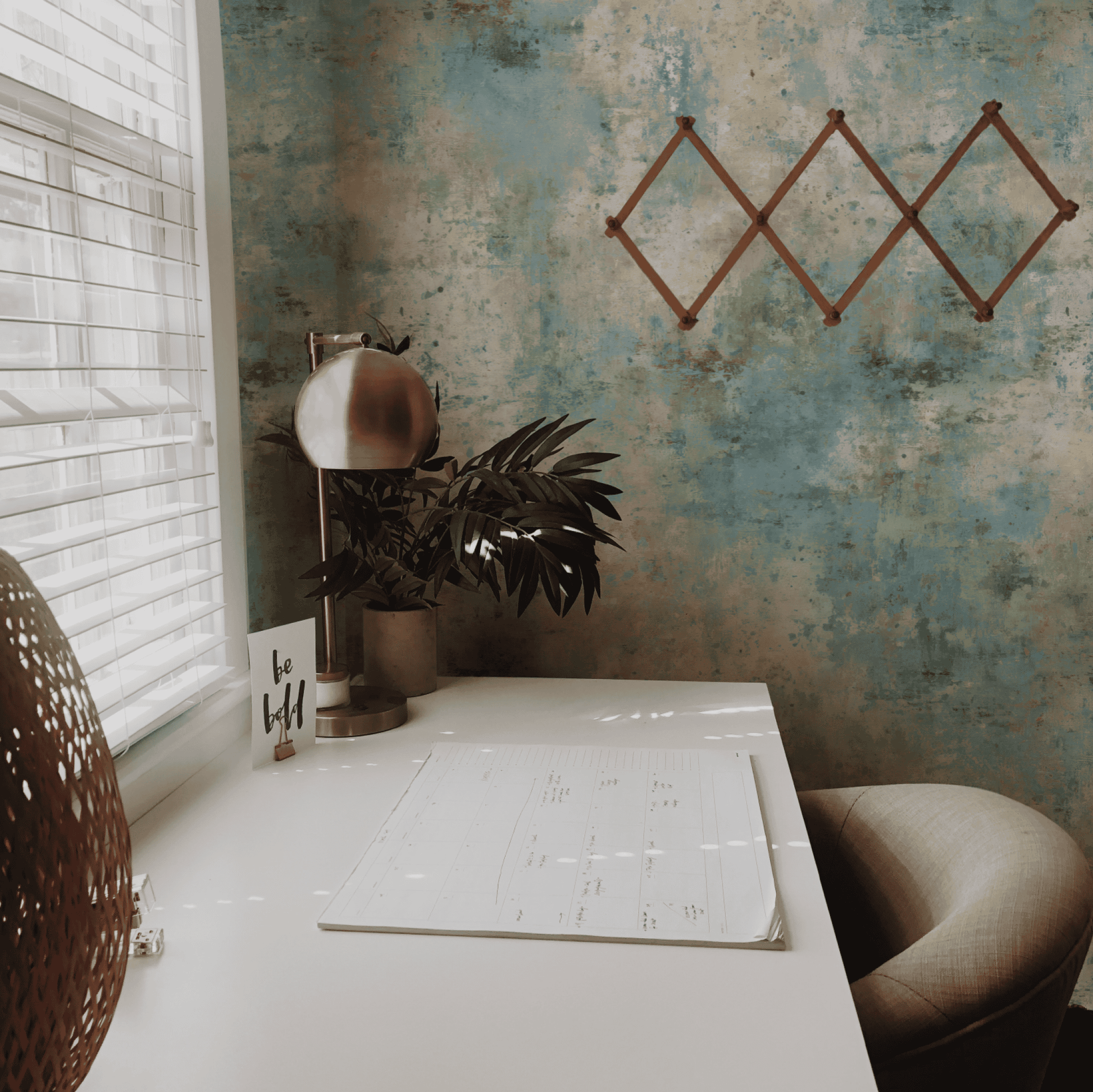 A minimalist home office desk with a lamp, plant, calendar, and 'be bold' card sits by a window with blinds; the Tranquil Mist Wallpaper in soft blue tones and an empty geometric rack create a calming ambiance.