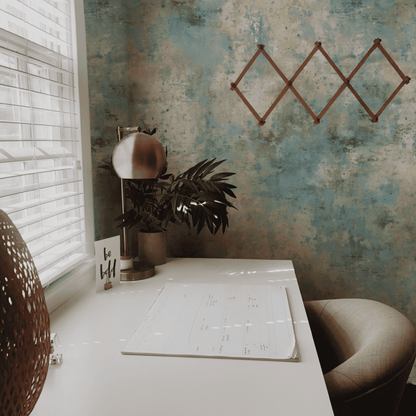 A minimalist home office desk with a lamp, plant, calendar, and 'be bold' card sits by a window with blinds; the Tranquil Mist Wallpaper in soft blue tones and an empty geometric rack create a calming ambiance.