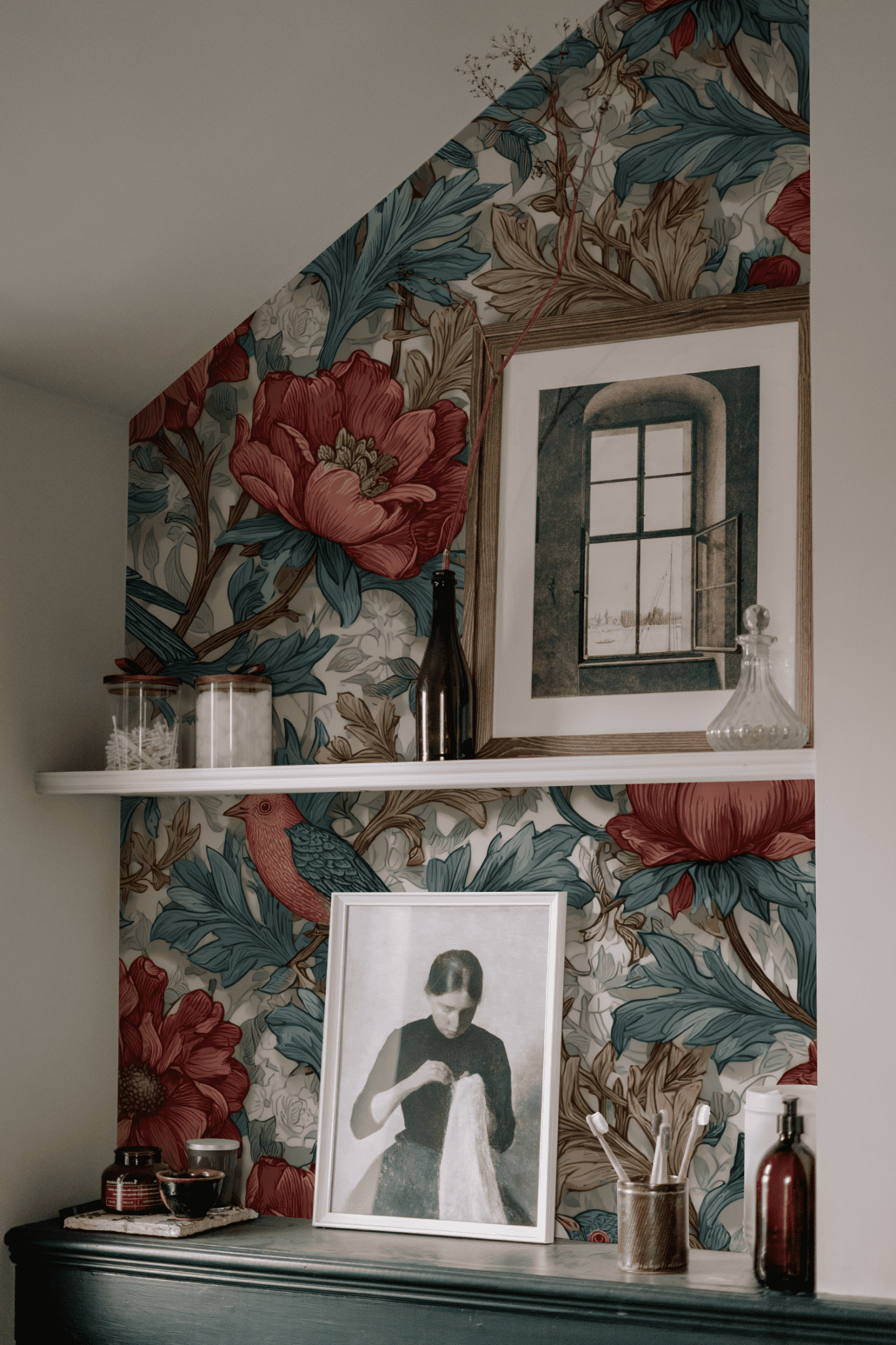 A wall with Chateau Bloom Wallpaper featuring bold crimson flowers, a white shelf displaying bottles and glassware, and framed art prints—including a vintage romance portrait—arranged on and above the shelf.