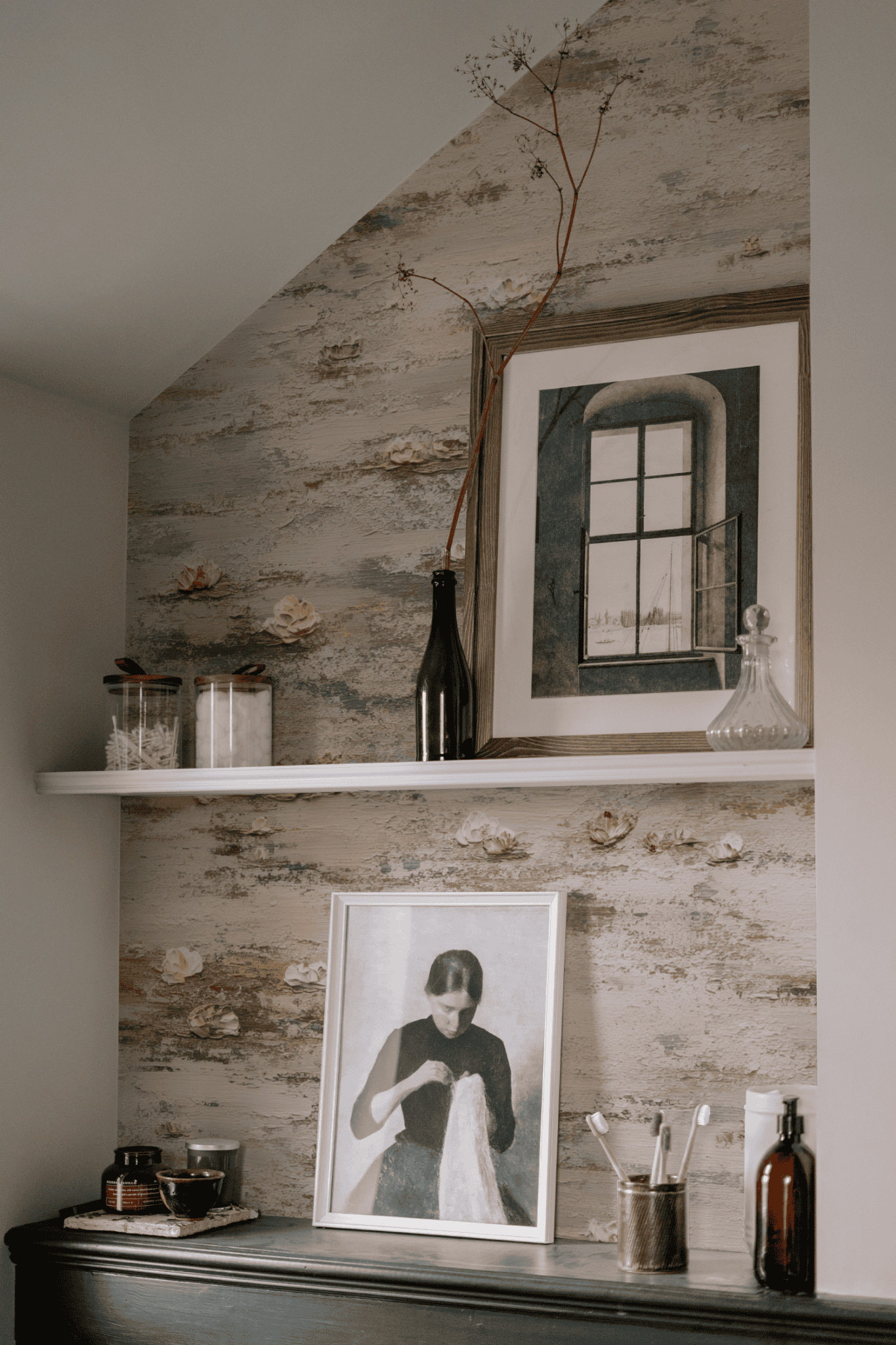A shelf displays jars, bottles, and framed art; beneath it, another photo leans against a rustic wall adorned with Serene Blossoms Wallpaper, while toiletries and small objects rest on a dark surface nearby, creating an elegant ambiance.