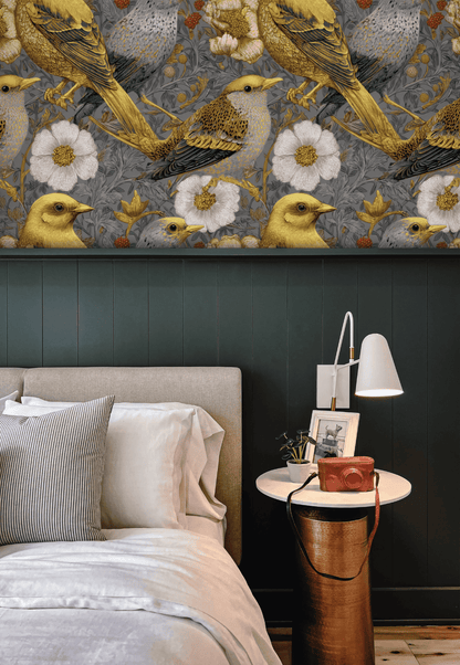 A cozy bedroom with a beige bed, striped pillow, green paneled wall, Golden Aviary Wallpaper featuring golden birds, and a round white side table topped with a lamp, camera, and books.