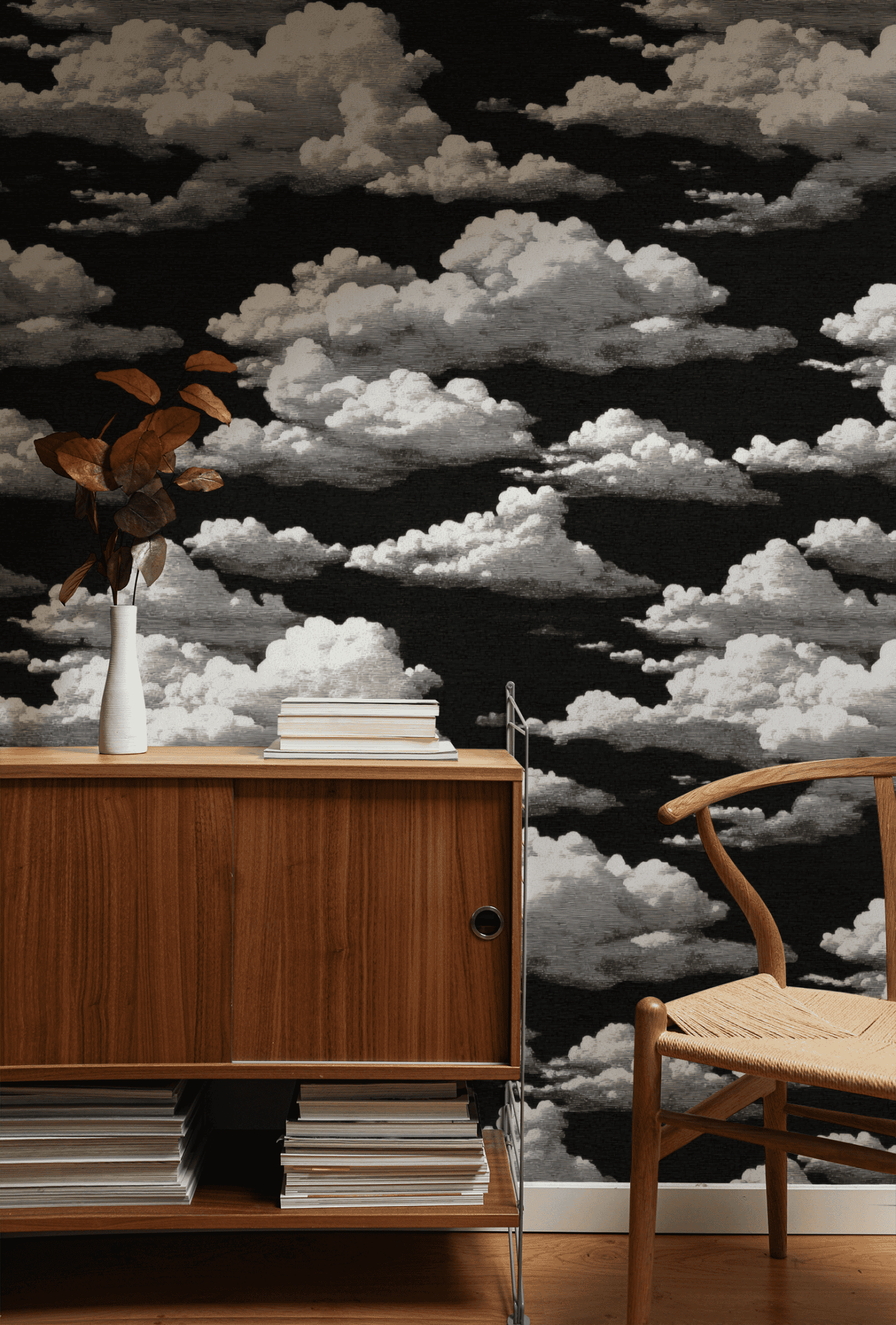 A wooden sideboard with stacked books and a white vase stands next to a woven chair; the background features Nimbus Dream Wallpaper, evoking a dreamy nightscape with white, fluffy clouds.