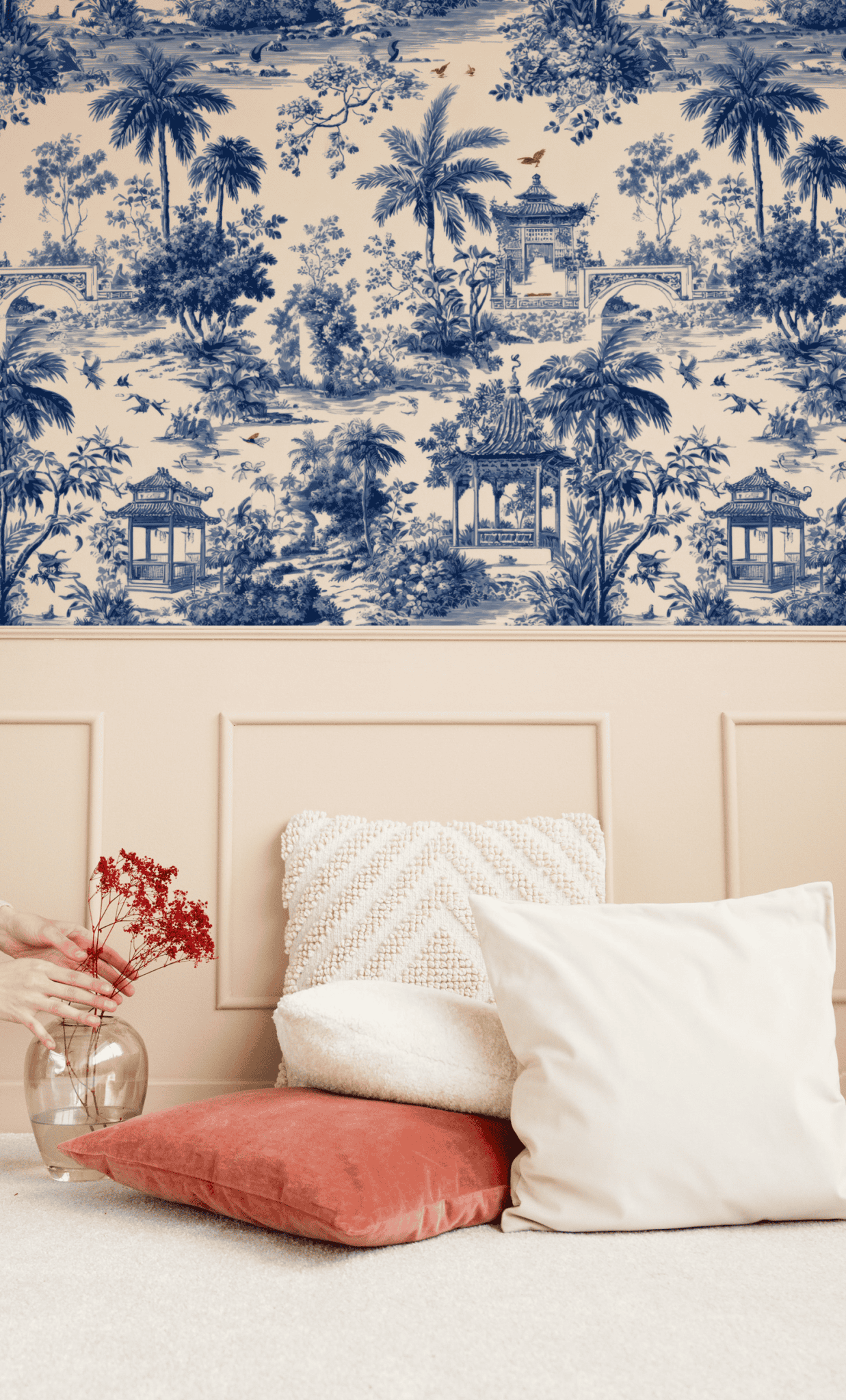A beige wall with Serene Pavilion Wallpaper in blue, a light pink wainscot, two white pillows, a pink cushion, and a hand placing red flowers in a glass vase on a soft light carpet.