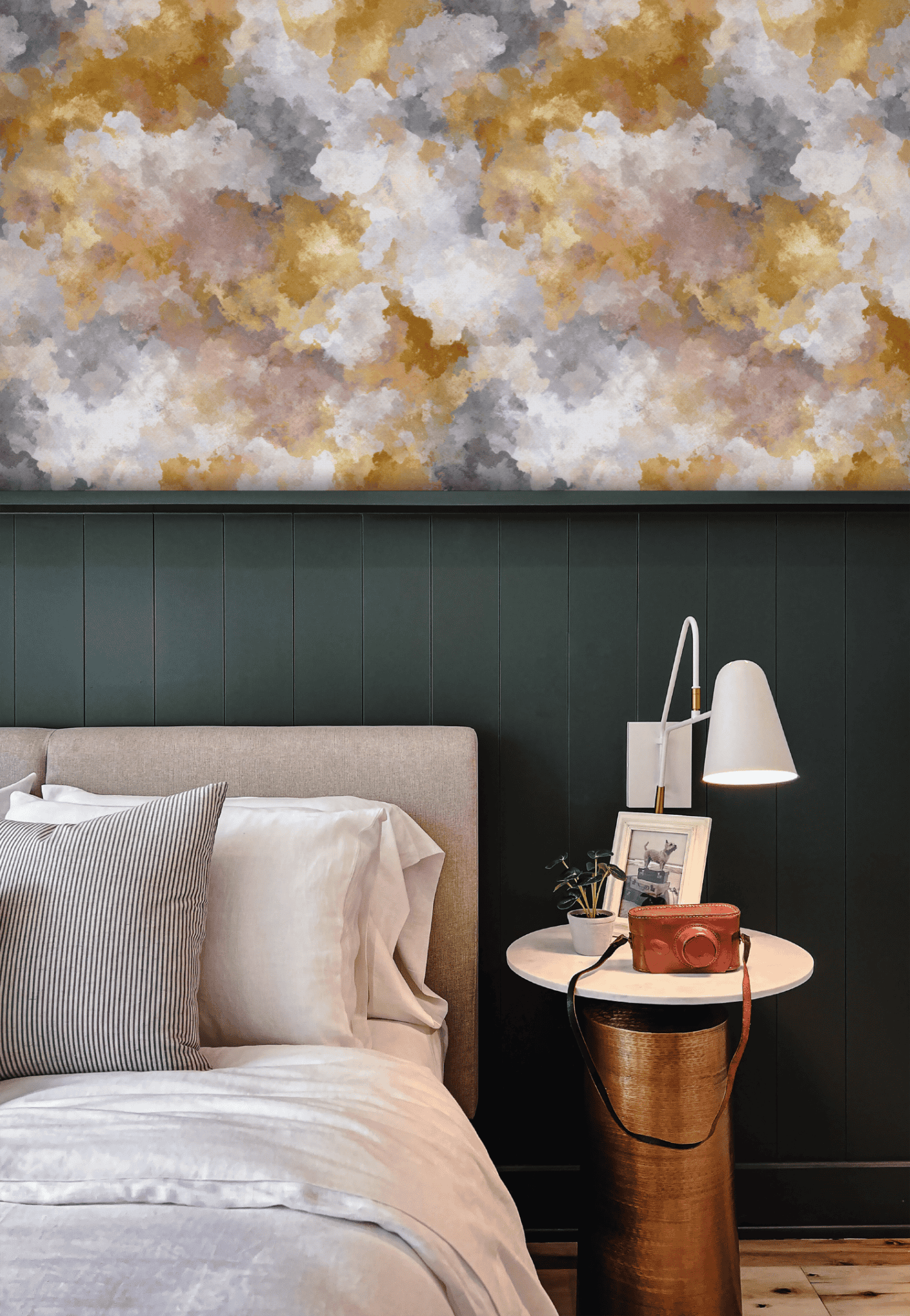 A neatly made bed with neutral bedding sits beside a round side table holding a white lamp, framed photos, and a camera, all set against a dark green paneled wall and Golden Serenity Wallpaper.