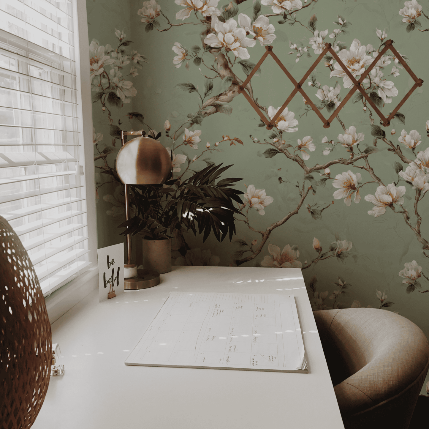 A desk with a planner, lamp, plant, and "be kind" sign sits by a window with blinds; the wall features Magnolia Whispers Wallpaper with green floral patterns and magnolia blossoms for a serene, tranquil atmosphere.