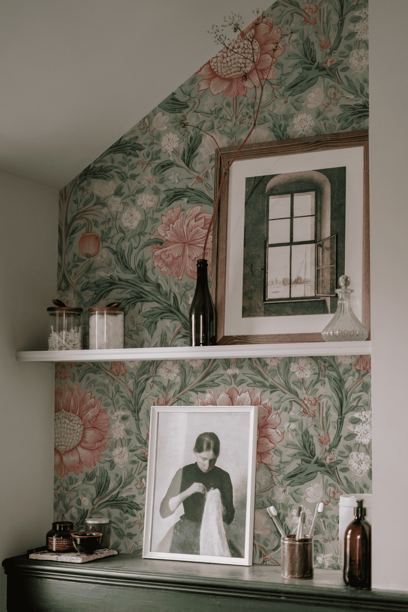 A shelf set against Eden Blossom Wallpaper, adorned with intricate foliage, holds jars, bottles, and two framed artworks—one of a window and the other depicting a woman with fabric.