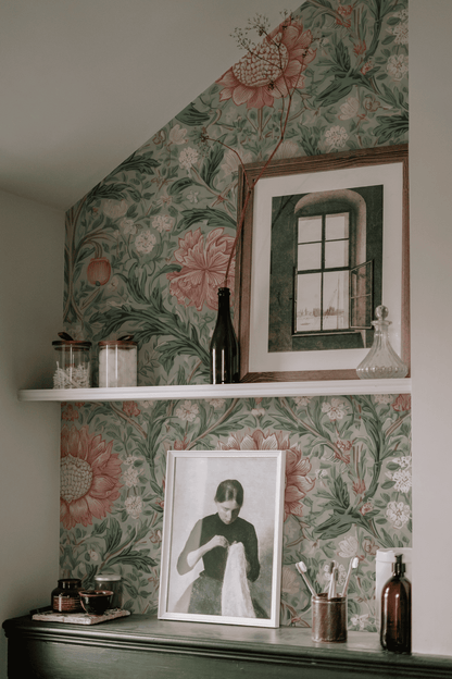A shelf set against Eden Blossom Wallpaper, adorned with intricate foliage, holds jars, bottles, and two framed artworks—one of a window and the other depicting a woman with fabric.
