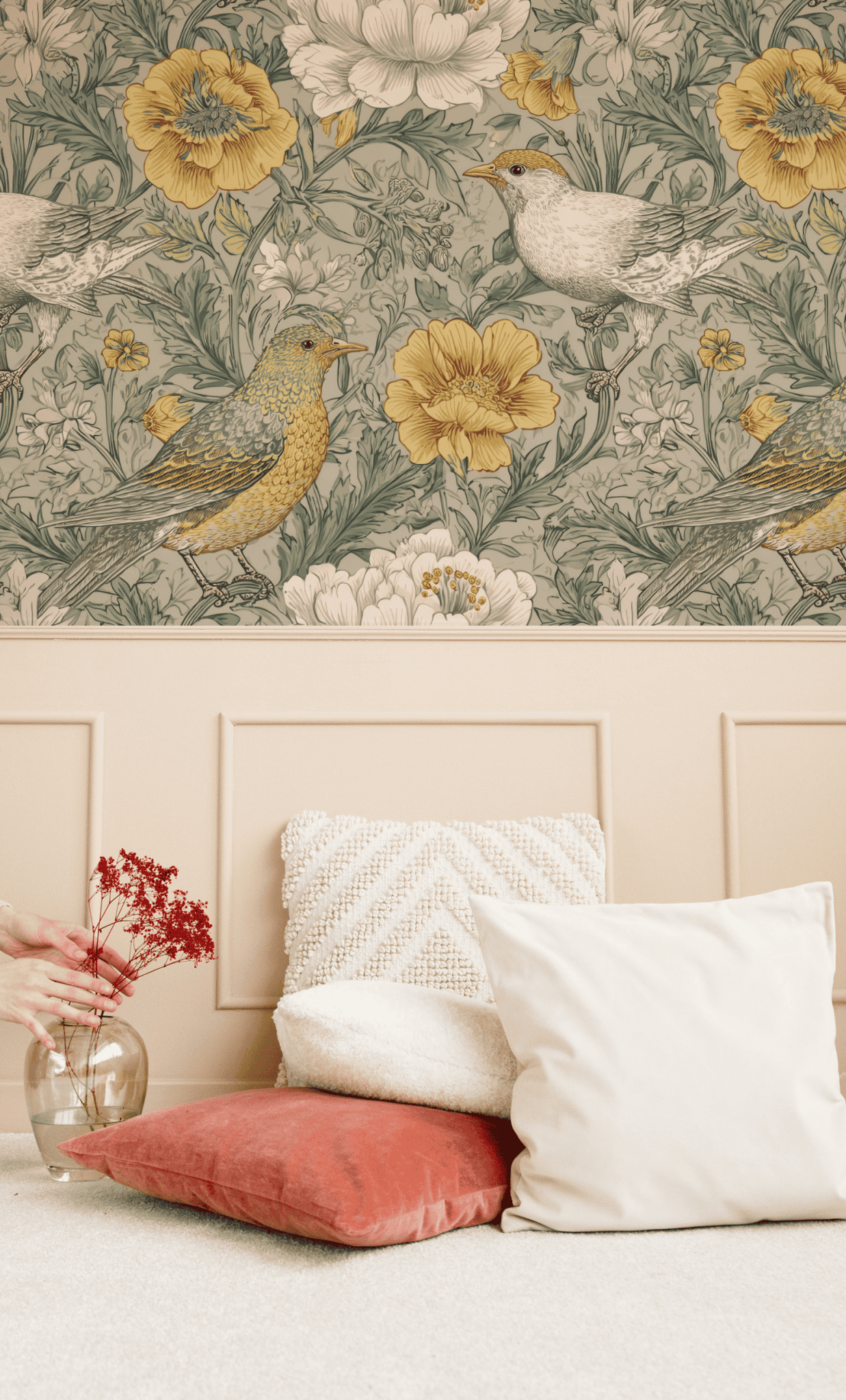 A wall with Meadow Whimsy Wallpaper featuring birds and florals above a beige wainscot, accented by white and rust-colored pillows and a glass vase of red flowers on the floor.