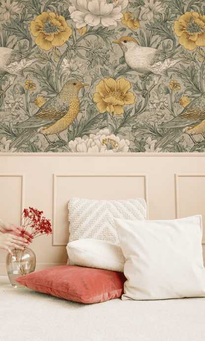 A wall with Meadow Whimsy Wallpaper featuring birds and florals above a beige wainscot, accented by white and rust-colored pillows and a glass vase of red flowers on the floor.