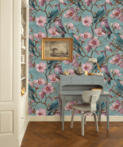 A blue desk and chair stand before a teal wall covered in Blossom Aviary Wallpaper, adorned with vibrant birds and blooming flowers; above the desk hangs a framed bird artwork.