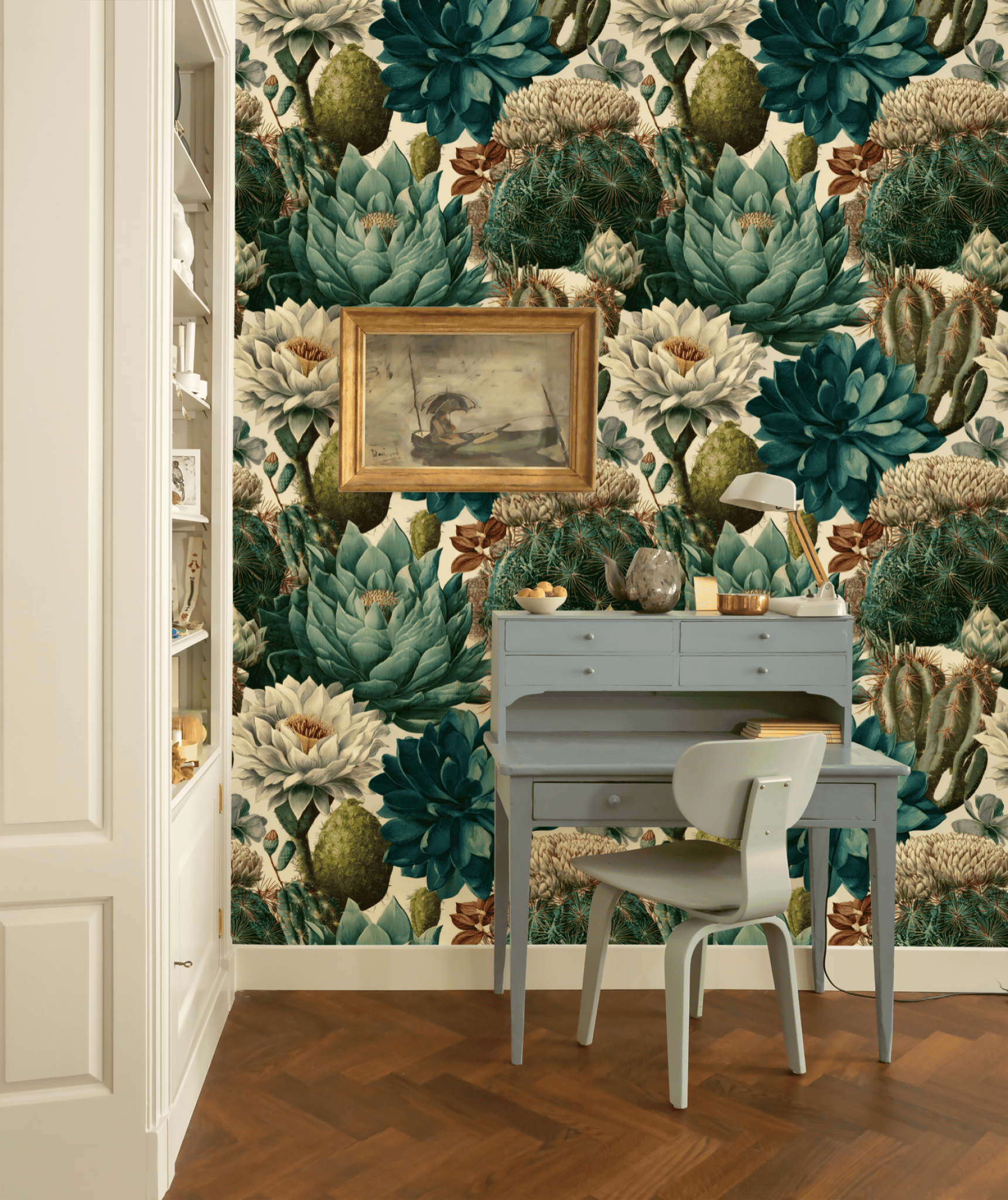 A small grey desk and chair sit against a wall adorned with Cactus Whisper Wallpaper’s bold green and cream florals with subtle cactus motifs; a framed painting hangs above the desk.