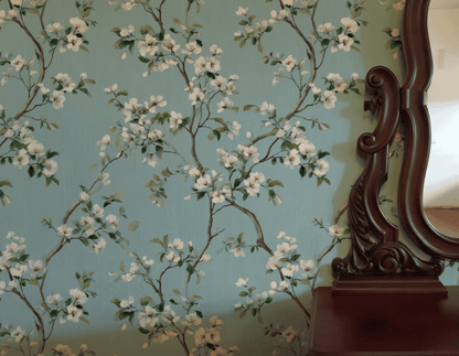 Blossom Dream Wallpaper features white blossoms on a light blue background, shown beside the ornately carved edge of a wooden mirror.