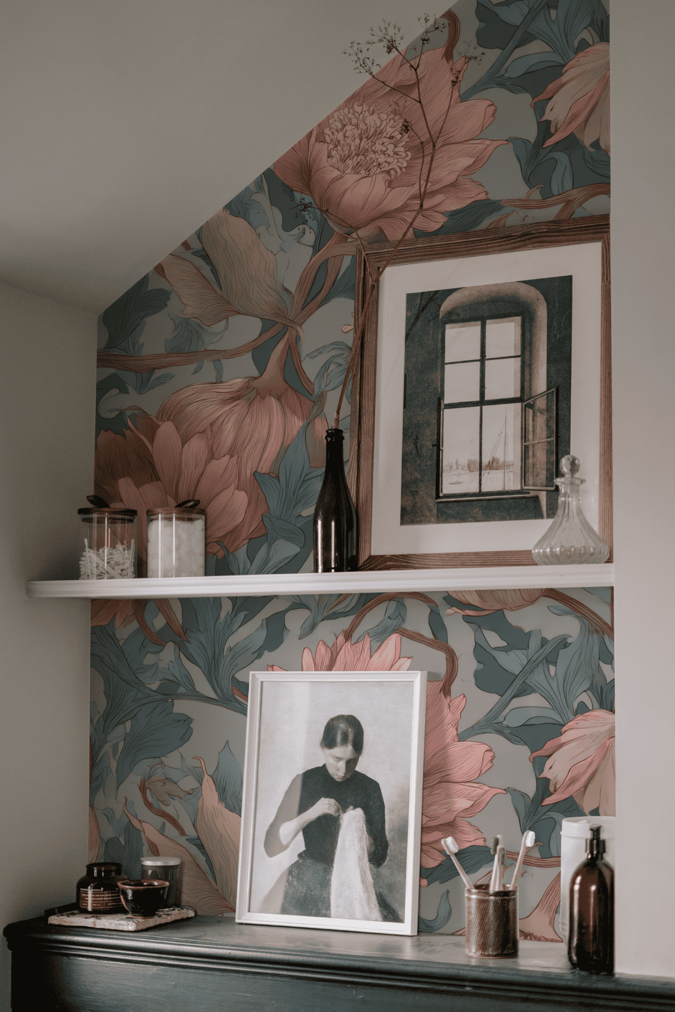 A shelf with glass jars, bottles, framed art, and photos stands against a wall adorned with elegant Petal Reverie Wallpaper in muted botanical tones.