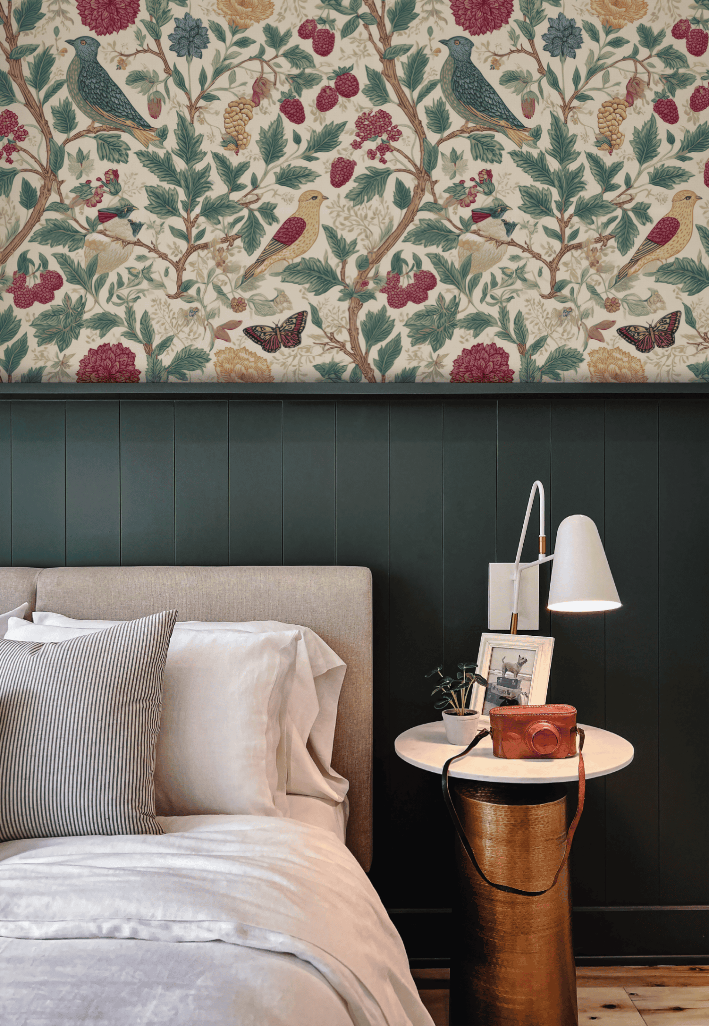 A bedroom with a beige upholstered bed, striped and white bedding, a round white nightstand with a lamp, camera, framed art, and elegant Garden Symphony Wallpaper featuring floral and bird designs.