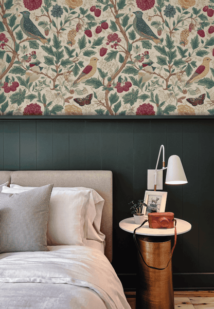 A bedroom with a beige upholstered bed, striped and white bedding, a round white nightstand with a lamp, camera, framed art, and elegant Garden Symphony Wallpaper featuring floral and bird designs.