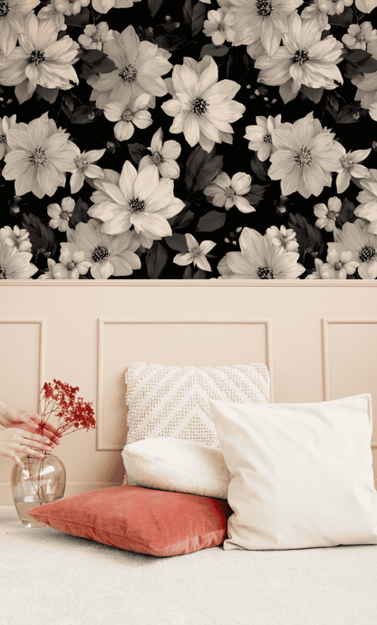 A hand places red flowers in a glass vase by cream and coral cushions on a beige carpet, with Monochrome Blossom Wallpaper adding elegance in the background.