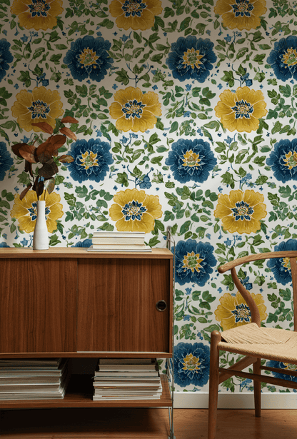 A wooden cabinet with stacked books and a vase of dried flowers stands against a wall decorated with Floral Harmony Wallpaper. A woven chair is partially visible on the right.