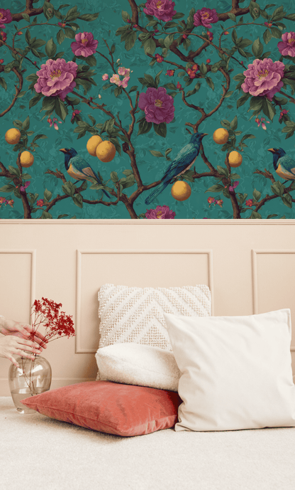 A hand arranges red dried flowers in a vase beside white and coral cushions on the floor, set against Eden Blooms Wallpaper with its floral and bird motifs in the background.