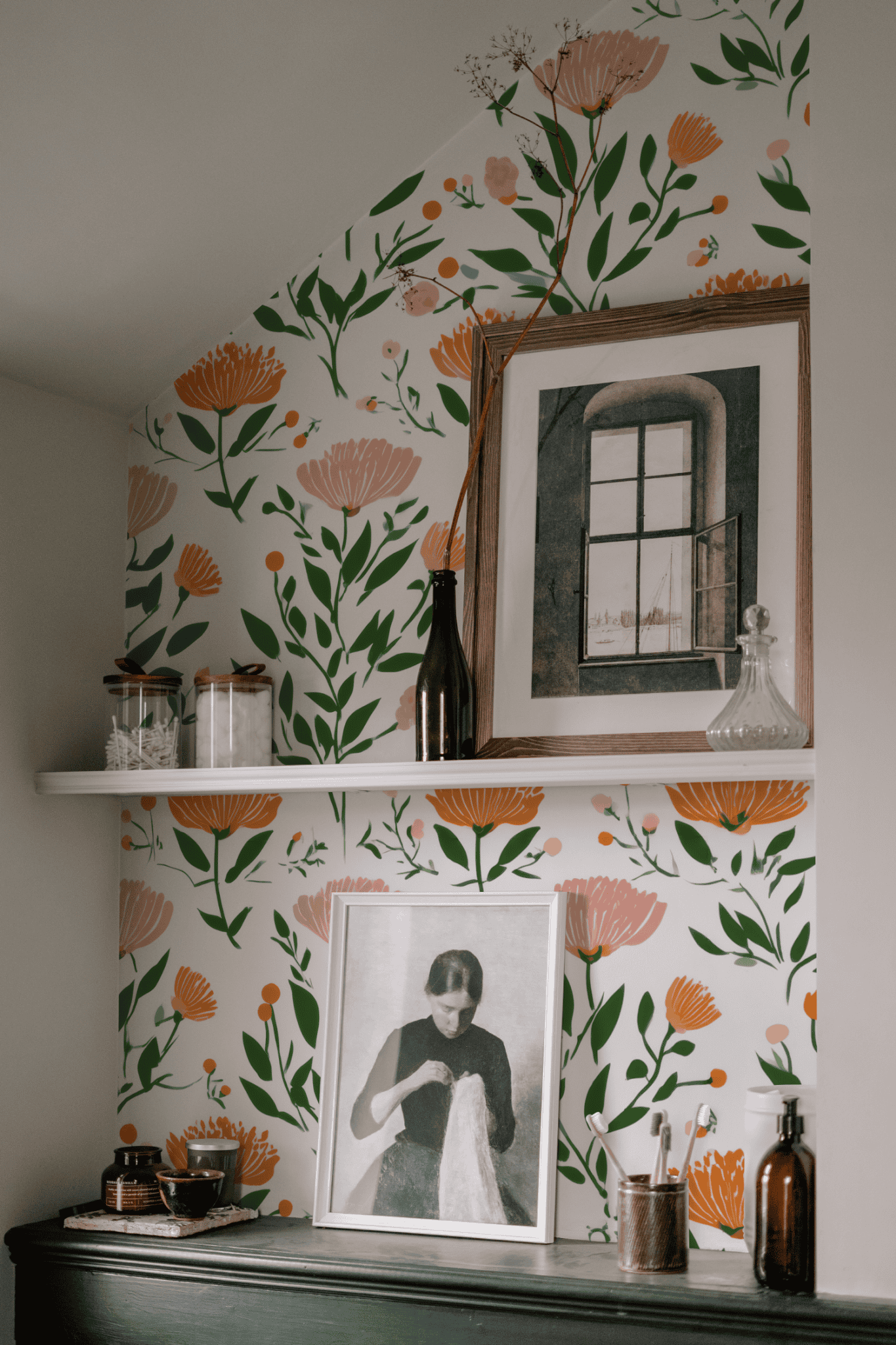 A shelf mounted on a Blossom Delight Wallpaper wall holds framed art, a glass decanter, jars, and vases; another framed photo leans against the wall below.