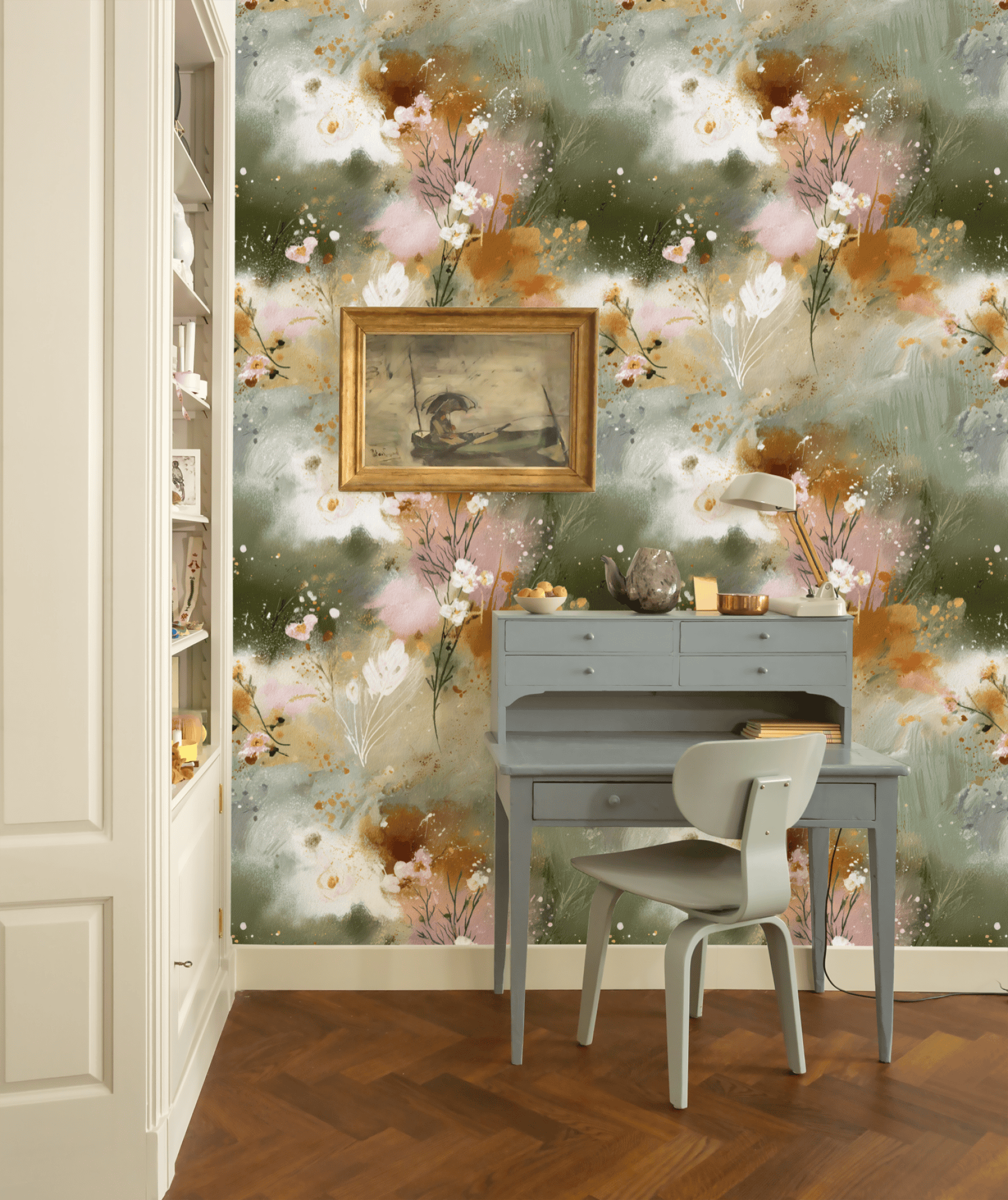 A small blue desk with a chair sits against Whispering Garden Wallpaper in muted greens. A framed painting hangs above, with a white lamp and books on the desk. Earthy-toned shelves to the left complete the nature-inspired look.