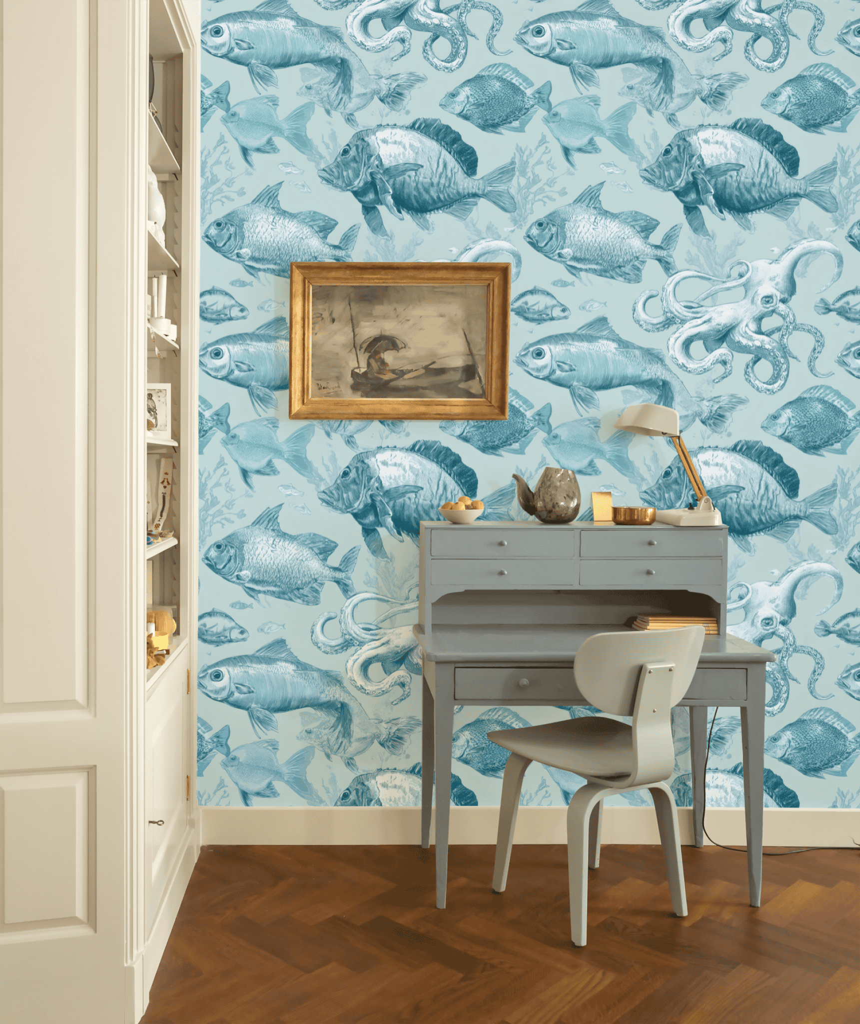 A small blue desk and chair sit against a wall with Aqua Nautica Wallpaper, featuring blue fish and sea creatures, complemented by a framed art piece and desk lamp above on a wooden floor.
