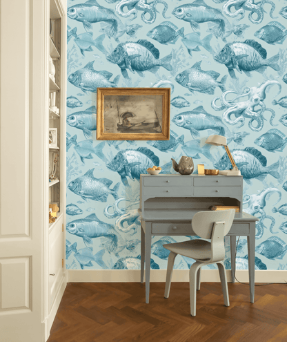 A small blue desk and chair sit against a wall with Aqua Nautica Wallpaper, featuring blue fish and sea creatures, complemented by a framed art piece and desk lamp above on a wooden floor.