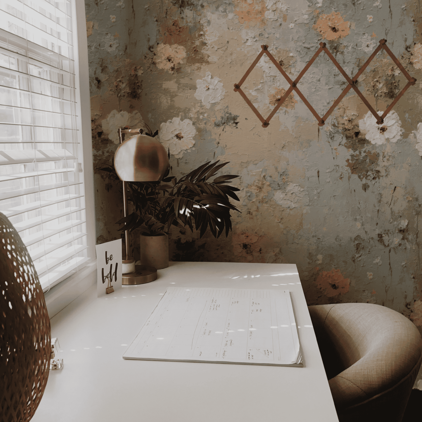 A white desk with a calendar, table lamp, and "be bold" card sits by a window with blinds in a room decorated with Whispering Blossoms Wallpaper and paired with a beige chair.