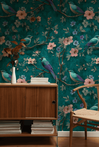 A wooden cabinet and chair are set against Serenade Bloom Wallpaper, featuring green tones, delicate flowers, illustrated teal birds, and pink blossoms. Books and a vase with dried leaves decorate the cabinet.