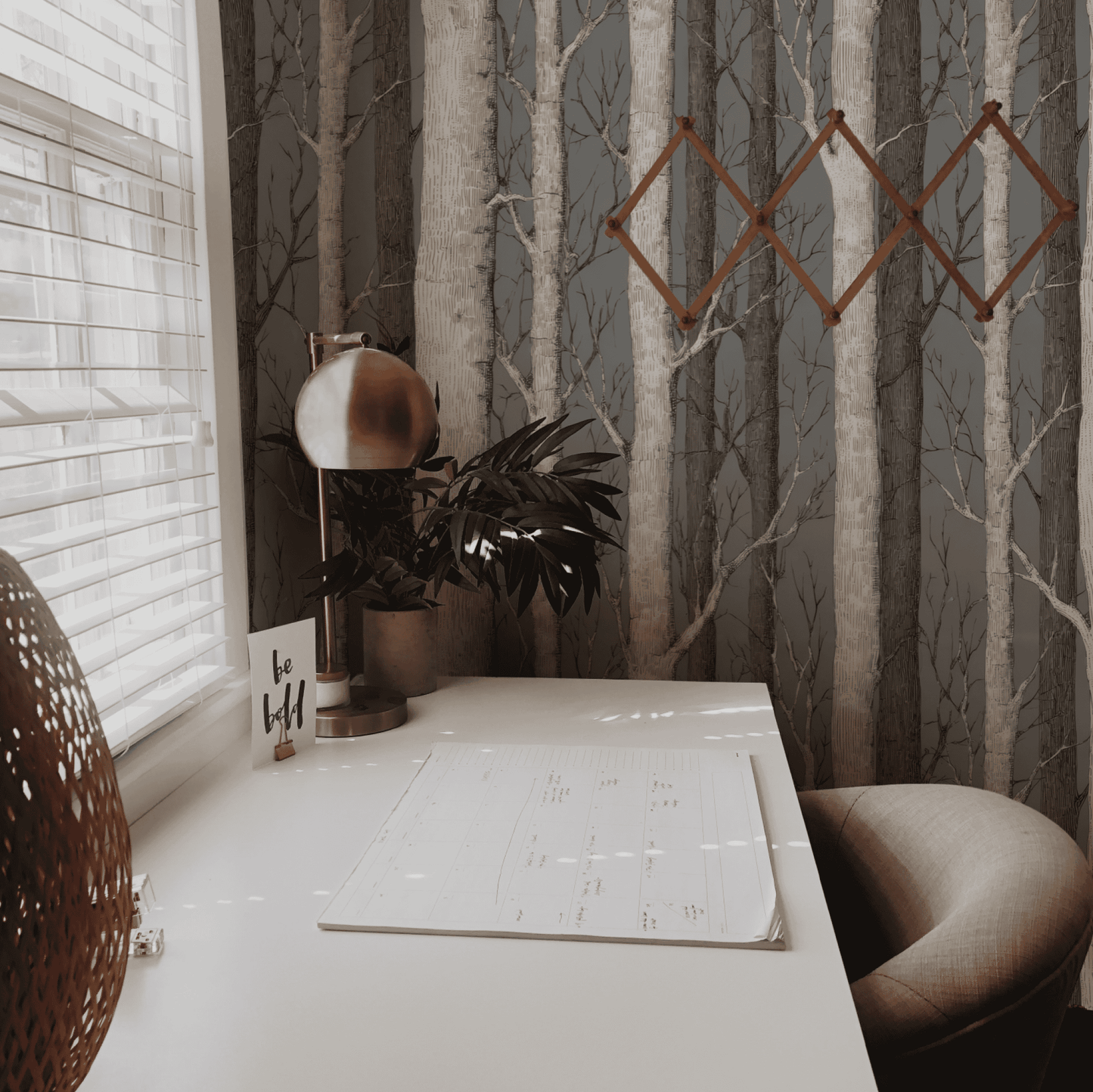 A minimal desk setup with a calendar, lamp, plant, and wall art sits by a window with blinds, complemented by Birch Grove Wallpaper for a touch of natural elegance.