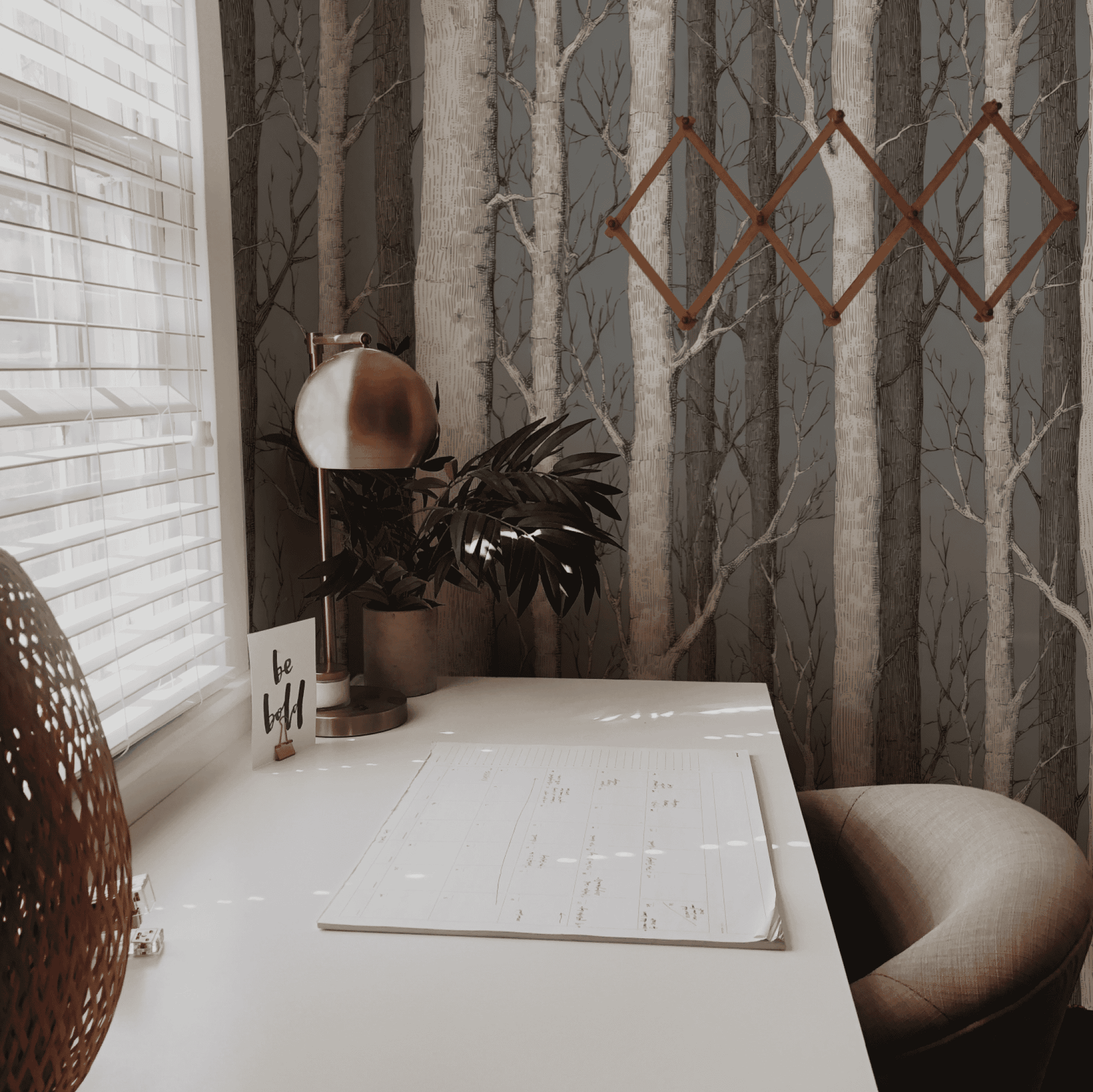 A minimal desk setup with a calendar, lamp, plant, and wall art sits by a window with blinds, complemented by Birch Grove Wallpaper for a touch of natural elegance.