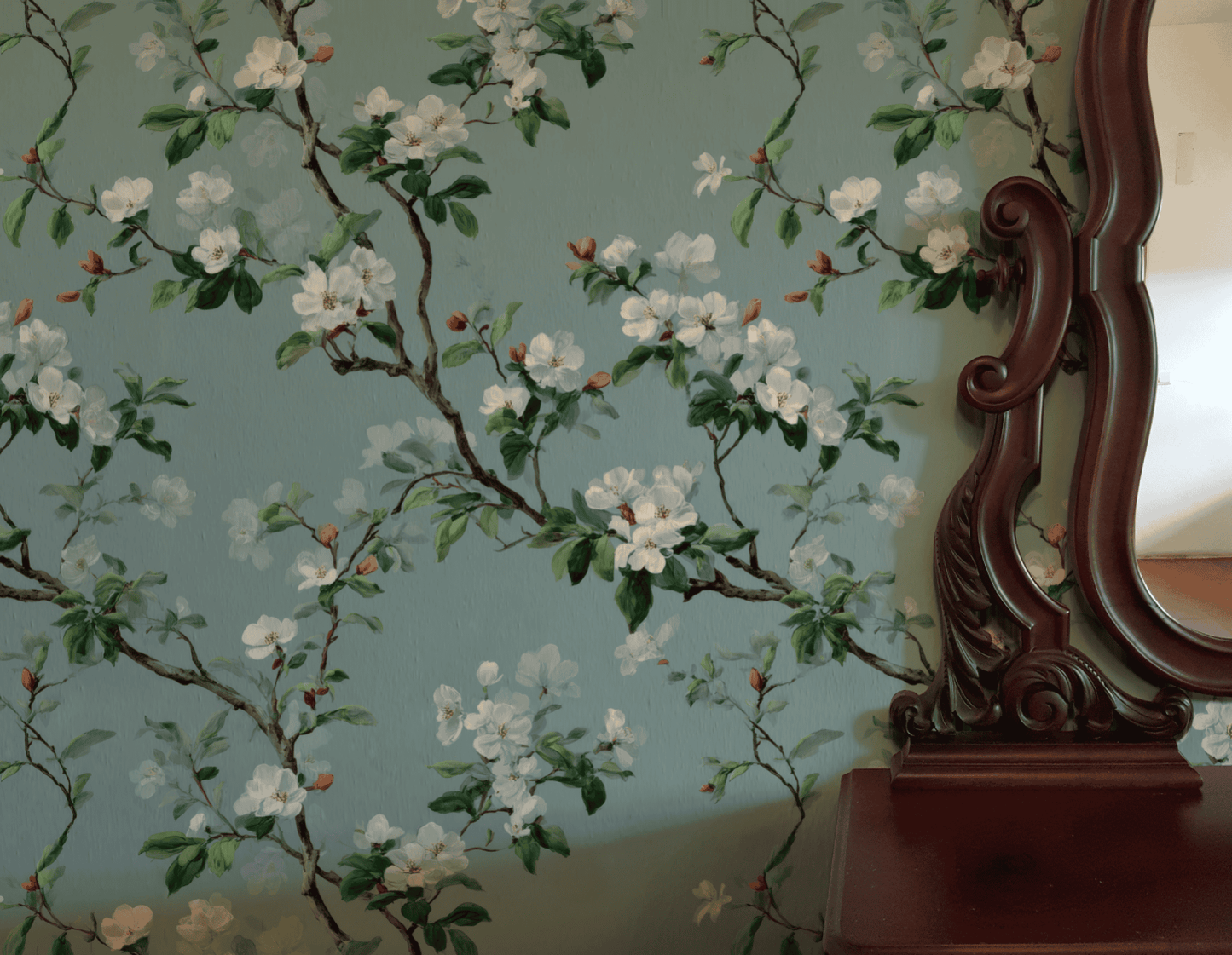 The Blossom Serenity Wallpaper, featuring white blossoms and green leaves on a soft teal background, brings tranquility to a space when paired with a vintage wooden mirror and a small wooden table.