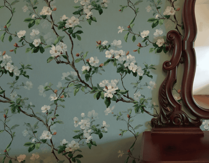 The Blossom Serenity Wallpaper, featuring white blossoms and green leaves on a soft teal background, brings tranquility to a space when paired with a vintage wooden mirror and a small wooden table.