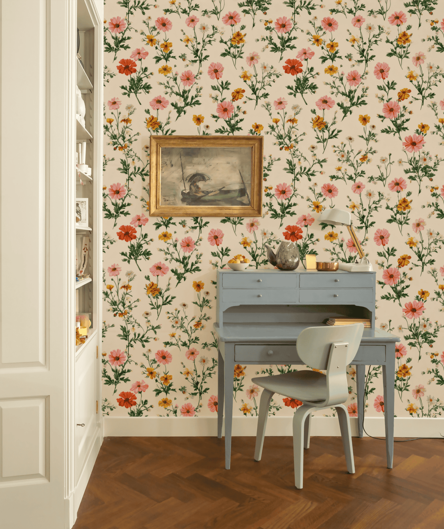 A small blue desk with a chair sits against a wall covered in Flora Charm Wallpaper; above hangs a bird painting, while a lamp and decor items rest on the desk.