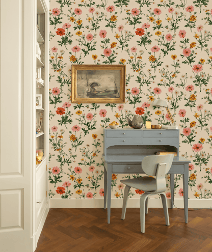 A small blue desk with a chair sits against a wall covered in Flora Charm Wallpaper; above hangs a bird painting, while a lamp and decor items rest on the desk.